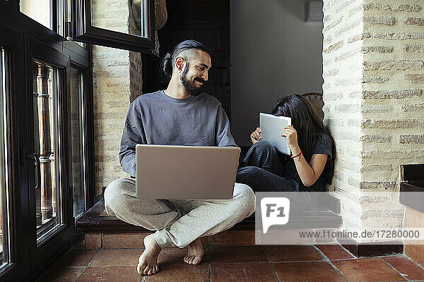 Smiling father looking at daughter hiding face with digital tablet at home