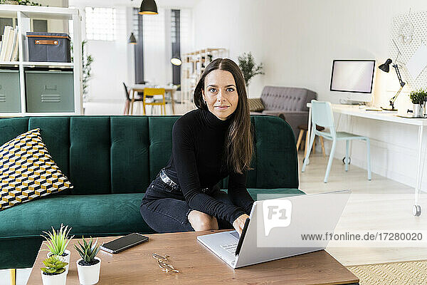 Beautiful young woman blogging through laptop while sitting on sofa in living room at loft apartment