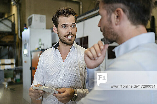 Junger Geschäftsmann mit Metallgegenstand im Gespräch mit einem Kollegen in einer modernen Fabrik