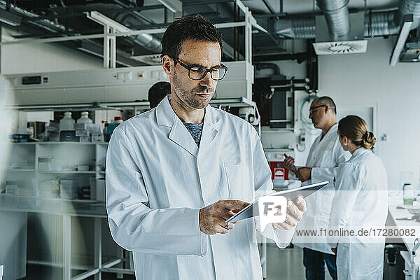 Wissenschaftlerin mit Brille  die ein digitales Tablet benutzt  während sie mit einem Mitarbeiter im Hintergrund in einem Labor steht