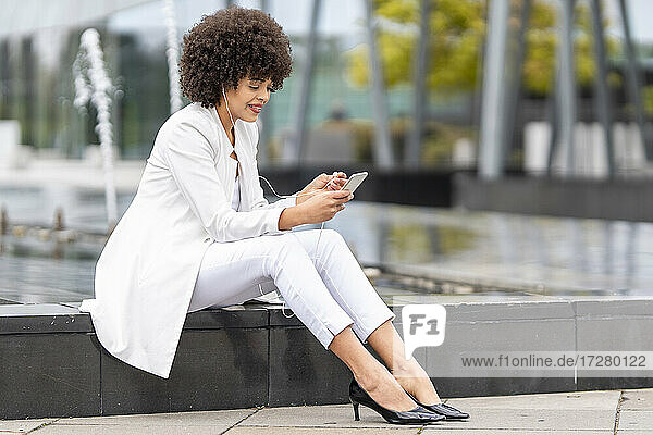 Geschäftsfrau mit In-Ear-Kopfhörern  die ein Mobiltelefon benutzt  während sie auf einer Bank im Freien sitzt