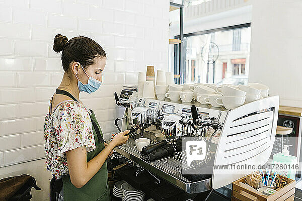 Junge Frau mit Gesichtsmaske beim Kaffeekochen in der Küche eines Cafés