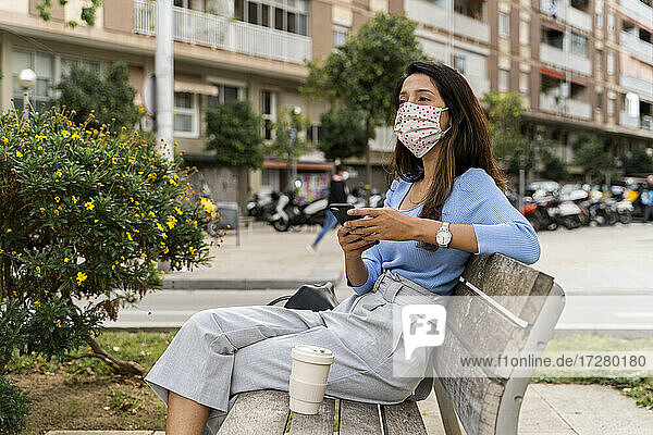 Frau  die mit ihrem Smartphone wegschaut  während sie auf einer Bank in der Stadt sitzt  während COVID-19