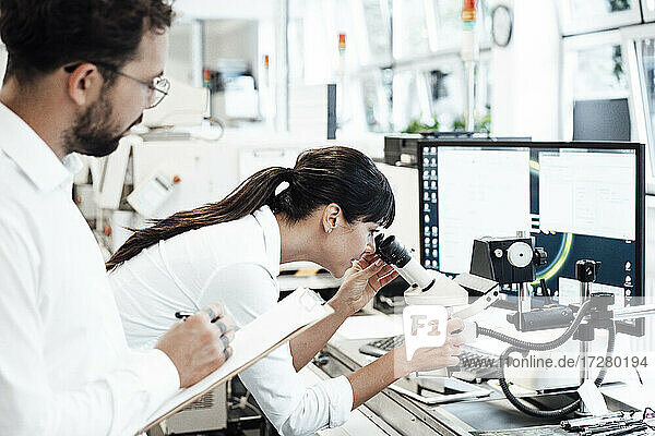 Female business professional looking through microscope while standing by male colleague at laboratory