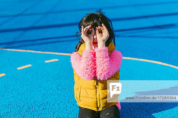 Girl forming binoculars with fingers while sitting at sports court