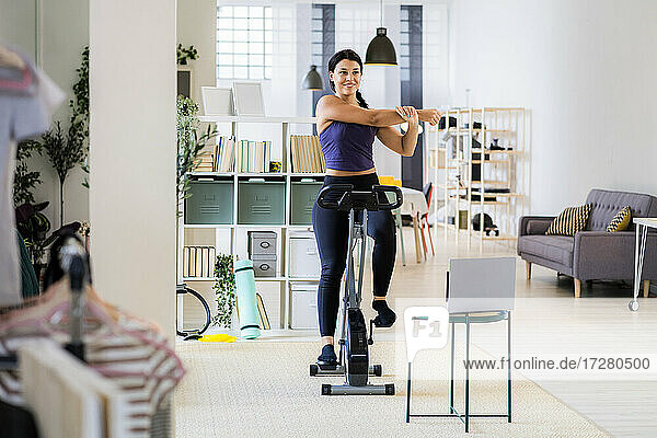Young female athlete smiling while doing stretching exercise sitting on exercise bike at home