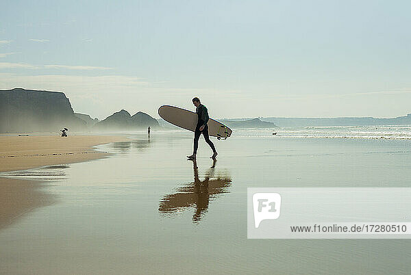 Spiegelung eines männlichen Teenagers  der mit einem Surfbrett gegen den Himmel am Strand läuft