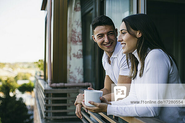 Lächelnder Mann sieht Frau mit Kaffeetasse auf dem Balkon stehend an