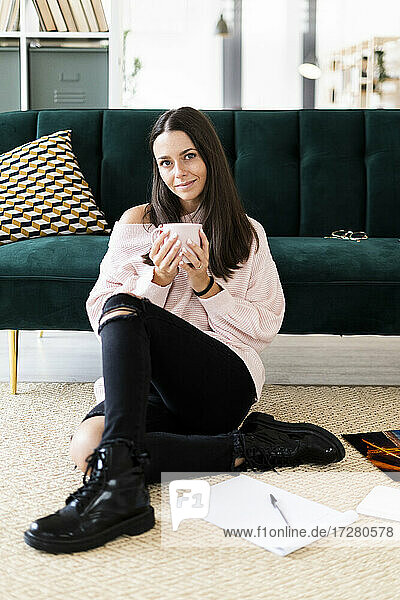Schöne junge Frau sitzt mit Kaffeetasse und Notizblöcken auf Teppich vor Sofa zu Hause