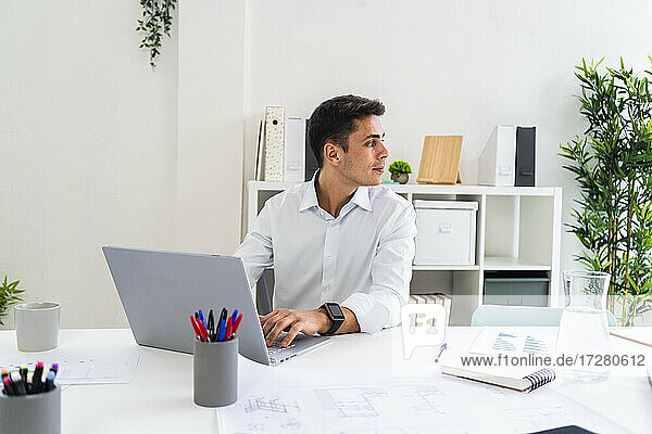 Männlicher Berufstätiger schaut weg  während er mit einem Laptop am Schreibtisch im Büro sitzt