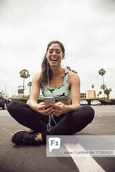 Junge Frau lacht  während sie auf der Straße sitzend ein Smartphone benutzt