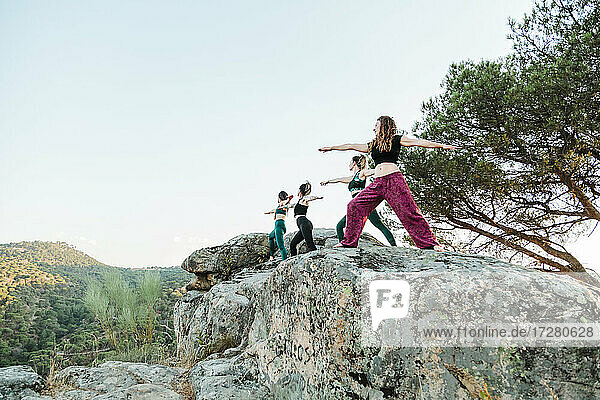 Female friends practicing yoga on rocks against clear sky during weekend