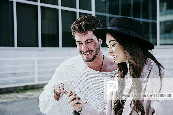 Lächelnde Frau und männlicher Partner schauen in der Stadt auf ihr Smartphone