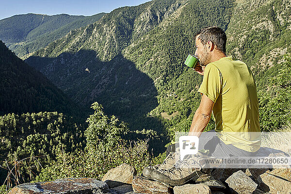 Mittlerer erwachsener Mann  der Kaffee trinkt  während er auf einem Stein im Wald an einem sonnigen Tag sitzt