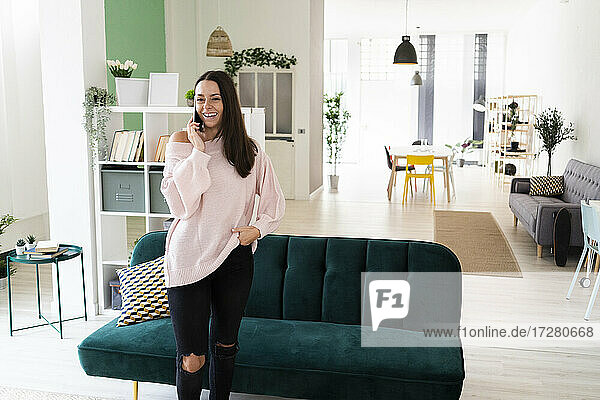 Happy beautiful young woman talking on mobile phone while standing in living room at loft apartment