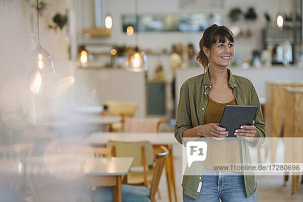 Lächelnde Unternehmerin  die wegschaut  während sie ein digitales Tablet in einem Café in der Hand hält