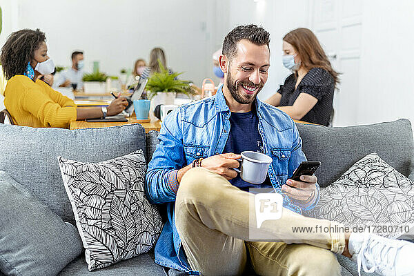 Smiling man holding coffee cup and mobile phone while sitting with coworker in background at office