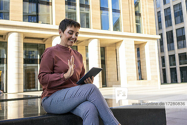 Woman waving hand to video call on digital tablet while sitting on bench against building