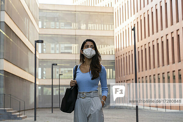 Geschäftsfrau mit Gesichtsmaske schaut weg  während sie vor einem Bürogebäude steht
