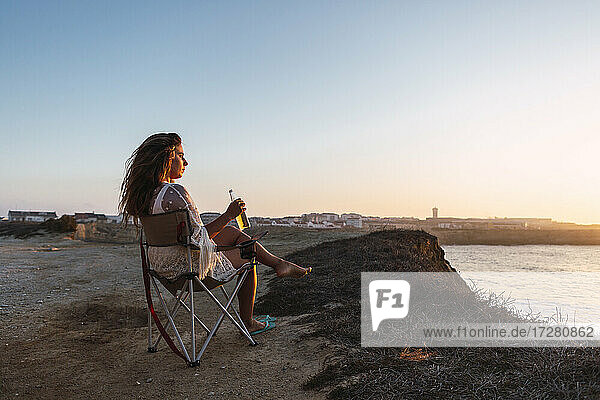 Frau trinkt Weißwein und sitzt bei Sonnenuntergang auf einem Stuhl am Strand