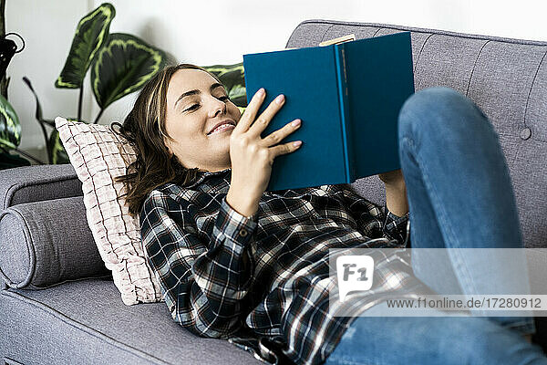 Lächelnde Frau liest ein Buch  während sie zu Hause auf dem Sofa liegt