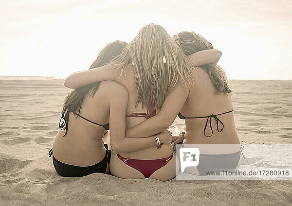 Female friends looking at view with arm around while sitting on beach during sunny day