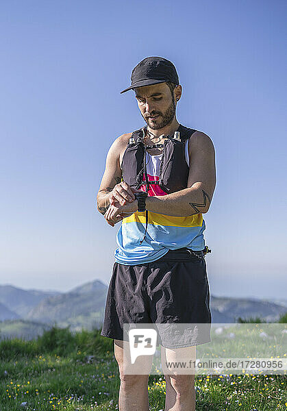 Mittlerer erwachsener Mann mit Mütze und Flaschengurt  der auf einem Berg stehend die Zeit auf der Uhr überprüft