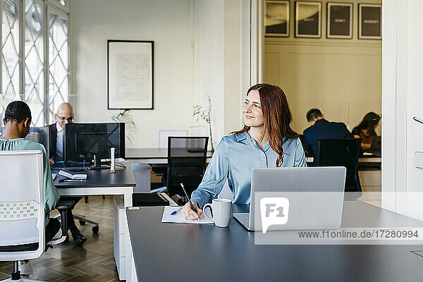 Lächelnde Geschäftsfrau beim Schreiben und Wegschauen im Büro