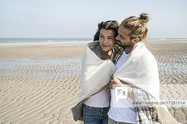 Romantisches junges Paar in Decke gehüllt am Strand stehend