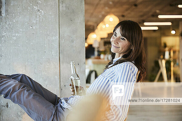 Lächelnde Frau  die wegschaut  während sie eine Bierflasche hält  die auf einem Stuhl zu Hause sitzt