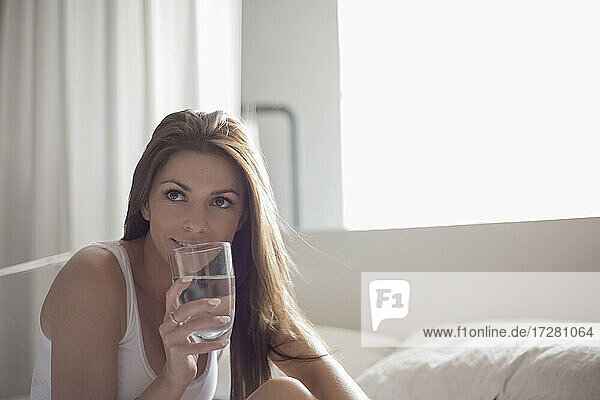 Junge Frau trinkt Wasser aus einem Glas im Schlafzimmer zu Hause