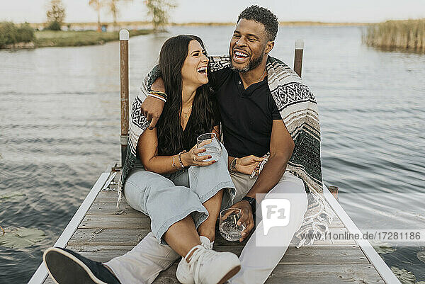 Happy couple drinking wine while sitting on pier against lake