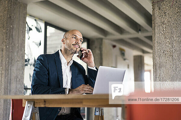 Businessman wearing suit talking on mobile phone while working on laptop at office