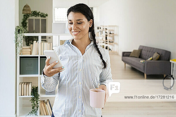 Lächelnde Frau  die eine Textnachricht auf einem Smartphone schreibt und eine Kaffeetasse hält  die zu Hause steht