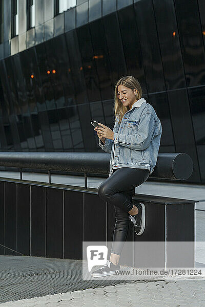 Lächelnde junge Frau  die ihr Smartphone benutzt  während sie sich an ein Geländer vor einem modernen Gebäude lehnt