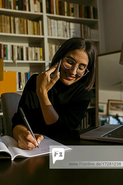 Lächelnde Geschäftsfrau  die mit ihrem Smartphone telefoniert  während sie in einem Buch schreibt  das zu Hause liegt