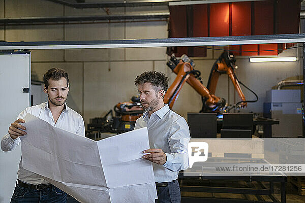 Team with blueprint having discussion while standing against robotic arm machine at factory