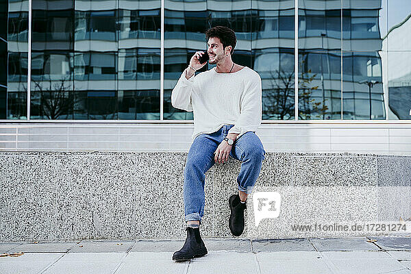 Smiling man talking on smart phone while sitting on retaining wall against building in city