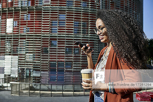 Lächelnde Frau  die eine Voicemail über ihr Smartphone sendet  während sie in der Stadt an einem sonnigen Tag steht