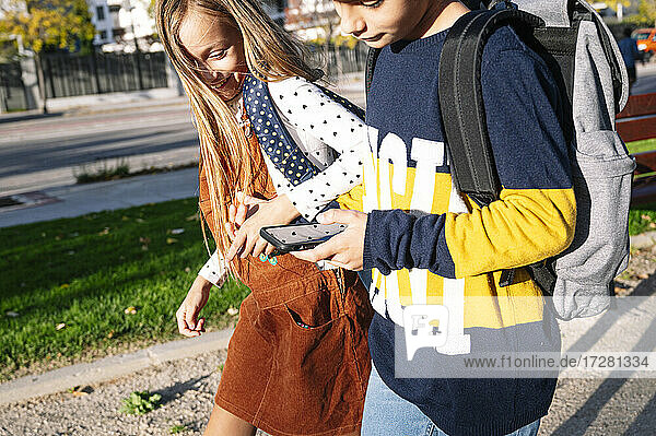 Geschwister halten sich an den Händen  während sie ihr Smartphone benutzen und in einem öffentlichen Park an einem sonnigen Tag spazieren gehen