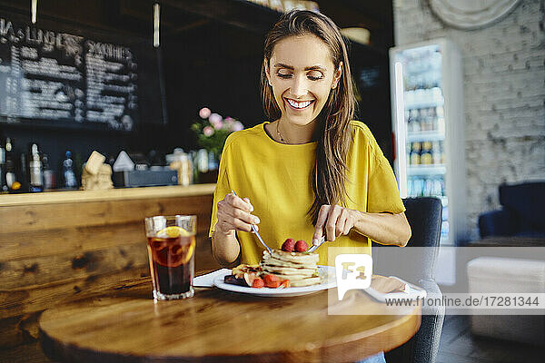 Smiling young female eating pancake while sitting at table in cafe