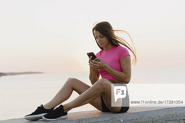Sportler  der ein Mobiltelefon benutzt  während er auf einer Betonwand am Meer sitzt