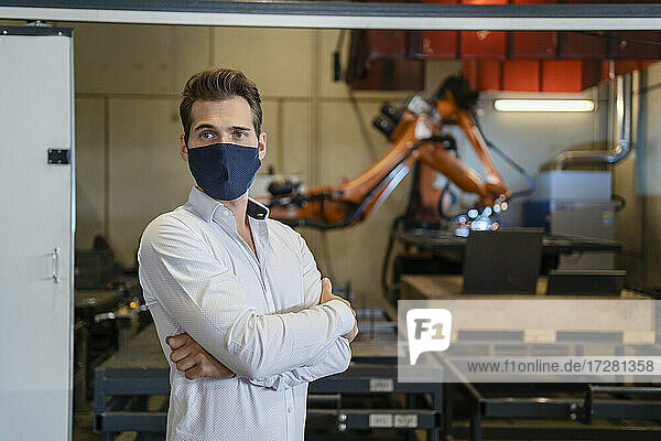 Businessman wearing face mask standing with arms crossed against robotic arm equipment at factory