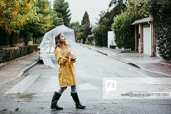 Smiling girl in raincoat holding umbrella while walking on road in city