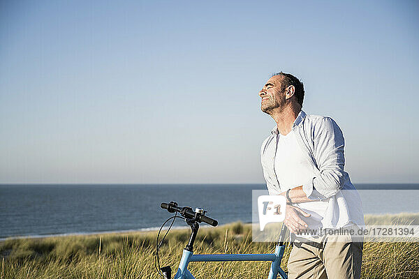 Mann mit geschlossenen Augen lehnt sich am Strand gegen den klaren Himmel auf ein Fahrrad