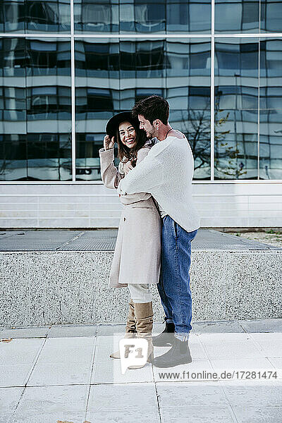 Lächelnder Mann  der seine Partnerin umarmt  während er an einem Gebäude in der Stadt steht