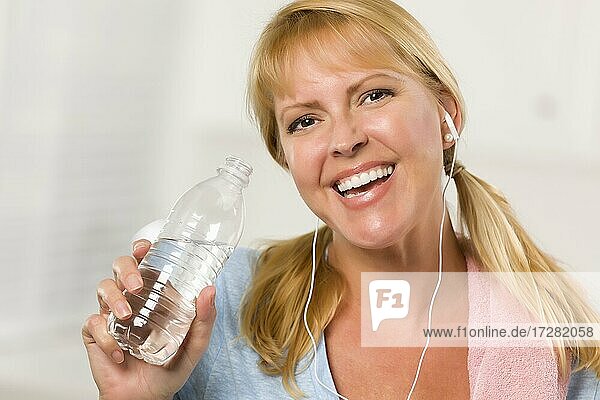 Hübsche blonde Frau mit Handtuch und Ohrhörer trinken aus Wasserflasche in ihrer Küche