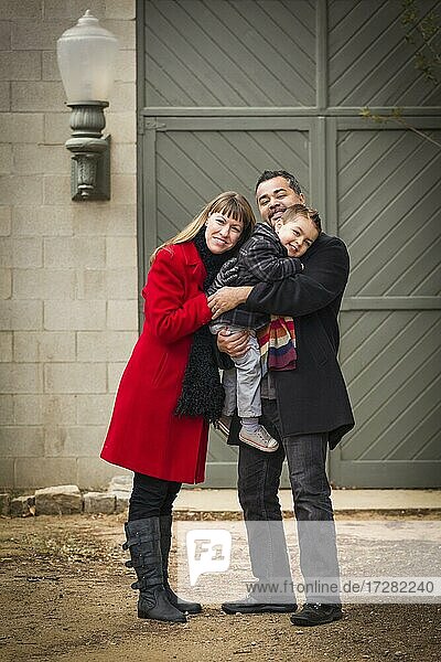 Young mixed-race couple in winter clothing hugging and kissing son in front of rustic building together