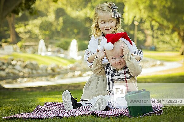 Süßes kleines Mädchen versucht  eine Weihnachtsmannmütze auf ihren widerwilligen kleinen Bruder im Freien im Park zu setzen