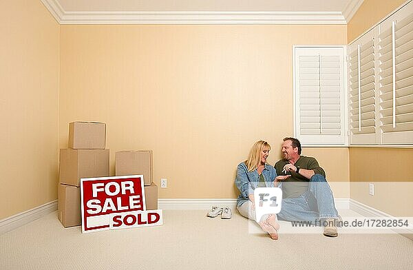 Aufgeregtes Paar mit neuen Schlüsseln  das sich auf dem Boden in der Nähe von Kisten und verkauften Immobilienschildern in einem leeren Raum entspannt